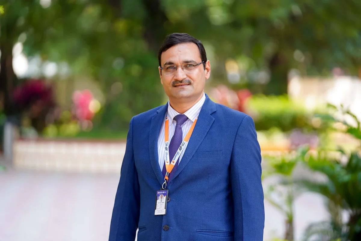 Professional male staff member in blue suit with Manipal lanyard standing in lush green campus garden setting