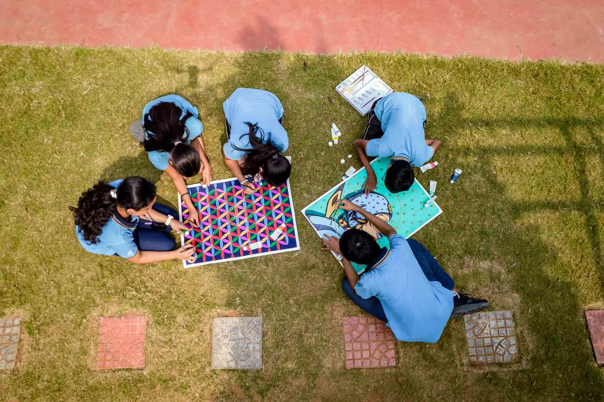 Students in blue uniforms sitting on grass working together on colorful geometric art projects during an outdoor activity